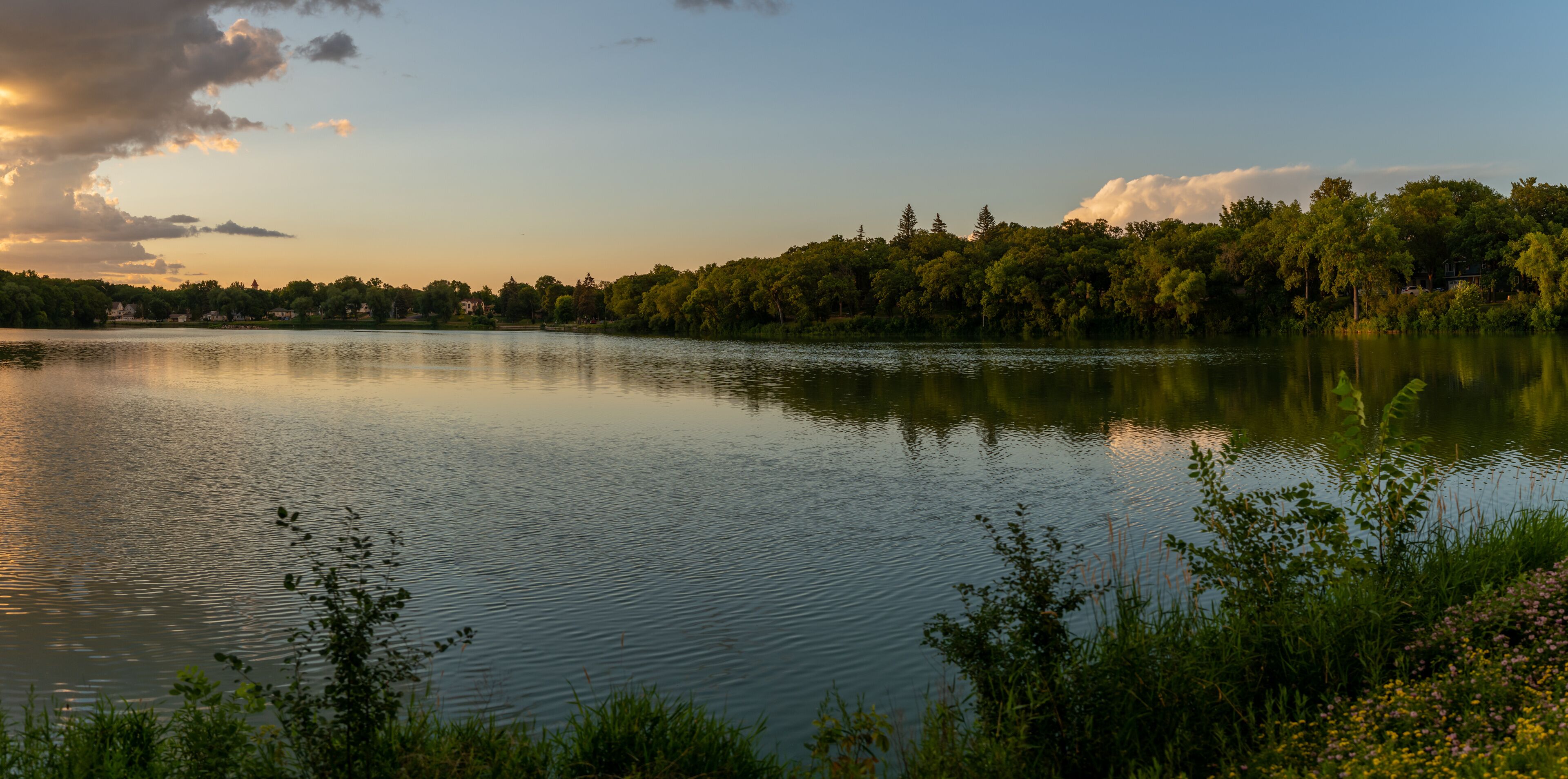 A calm lake with a forested shoreline and a few houses in the background.