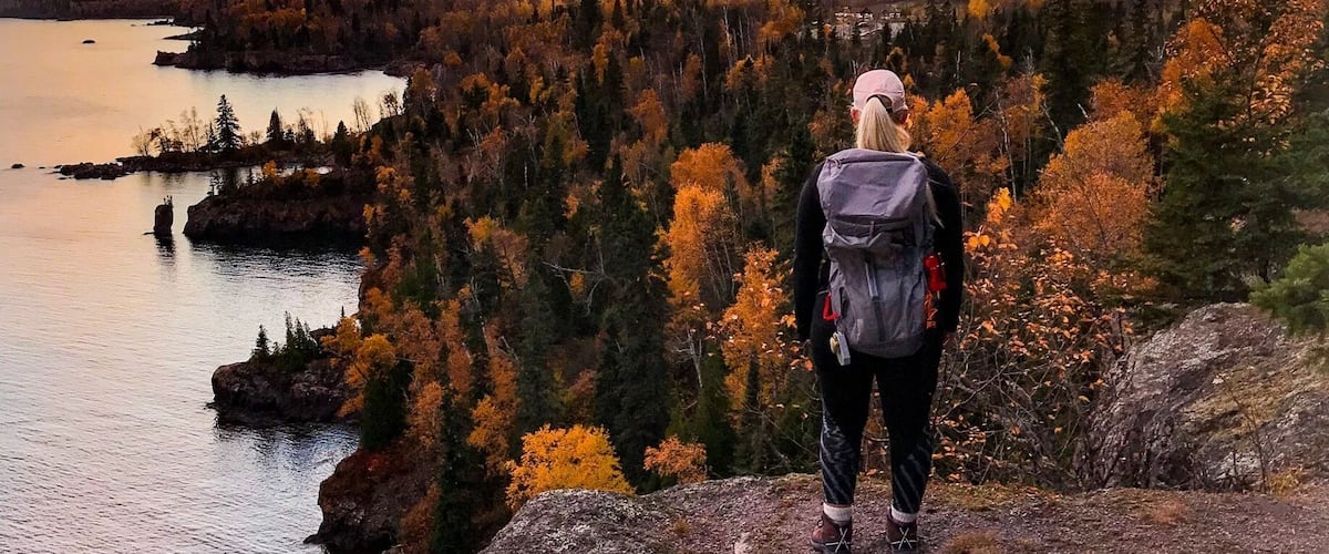 Un excursionista en un saliente rocoso contemplando un lago y un bosque al atardecer.