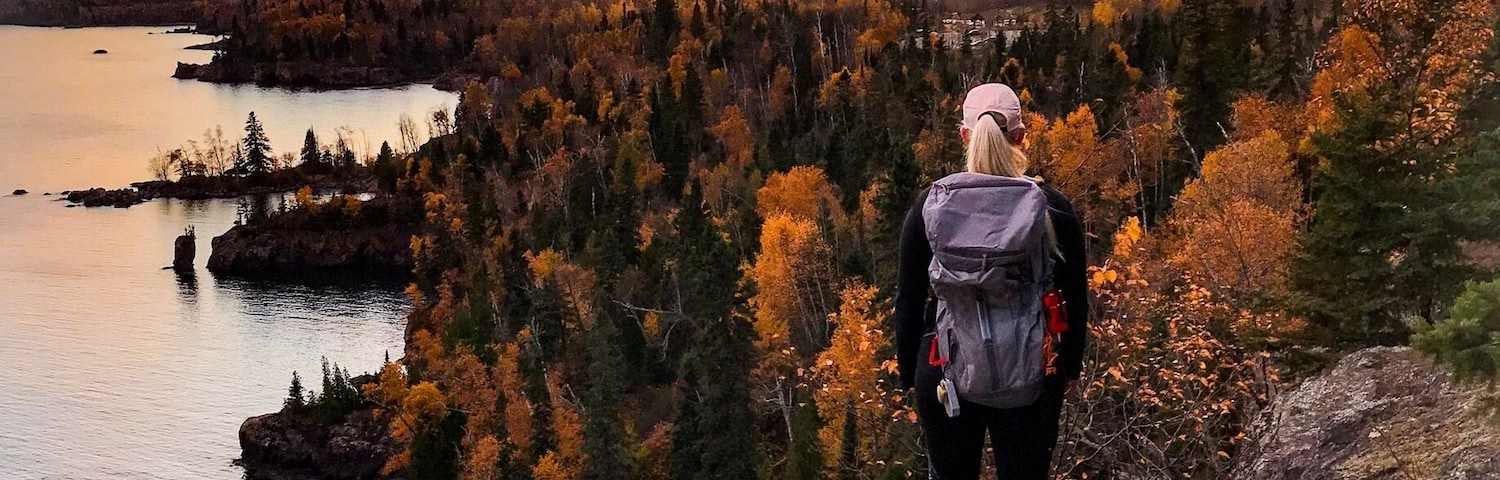 Un excursionista en un saliente rocoso contemplando un lago y un bosque al atardecer.