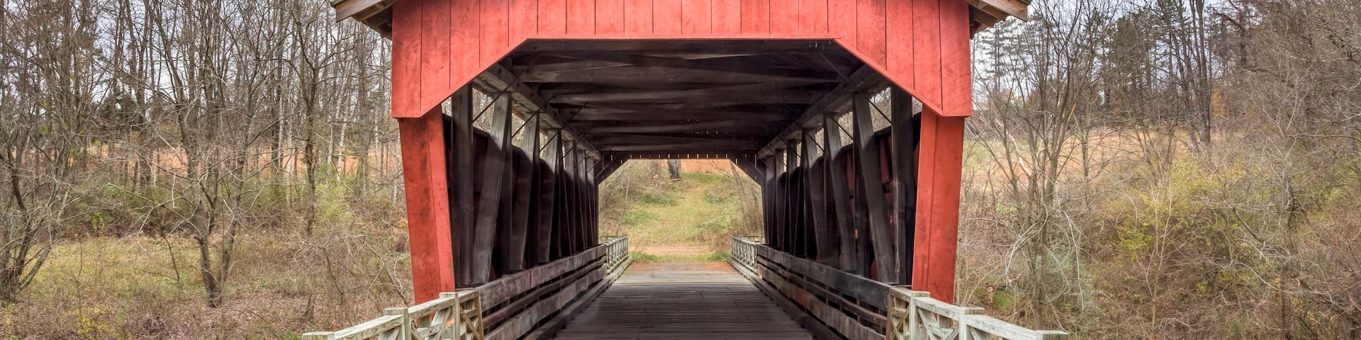 A wooden covered bridge with a red roof, surrounded by trees and grassy areas.