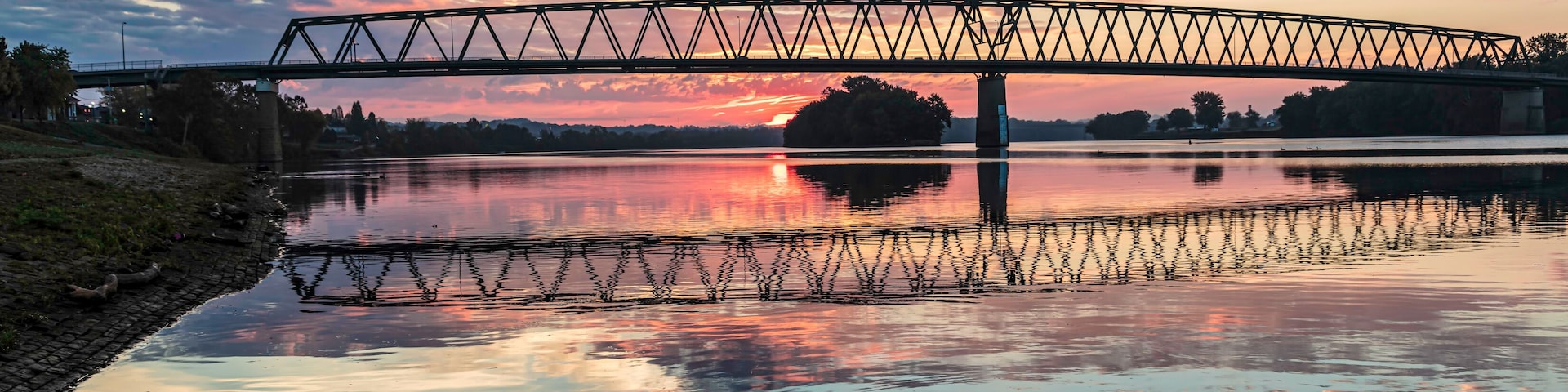 Un atardecer sobre un río tranquilo con un puente y una pequeña isla a lo lejos.