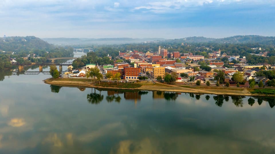 A town by a calm river with a bridge, historic buildings, and a hilly landscape in the background.