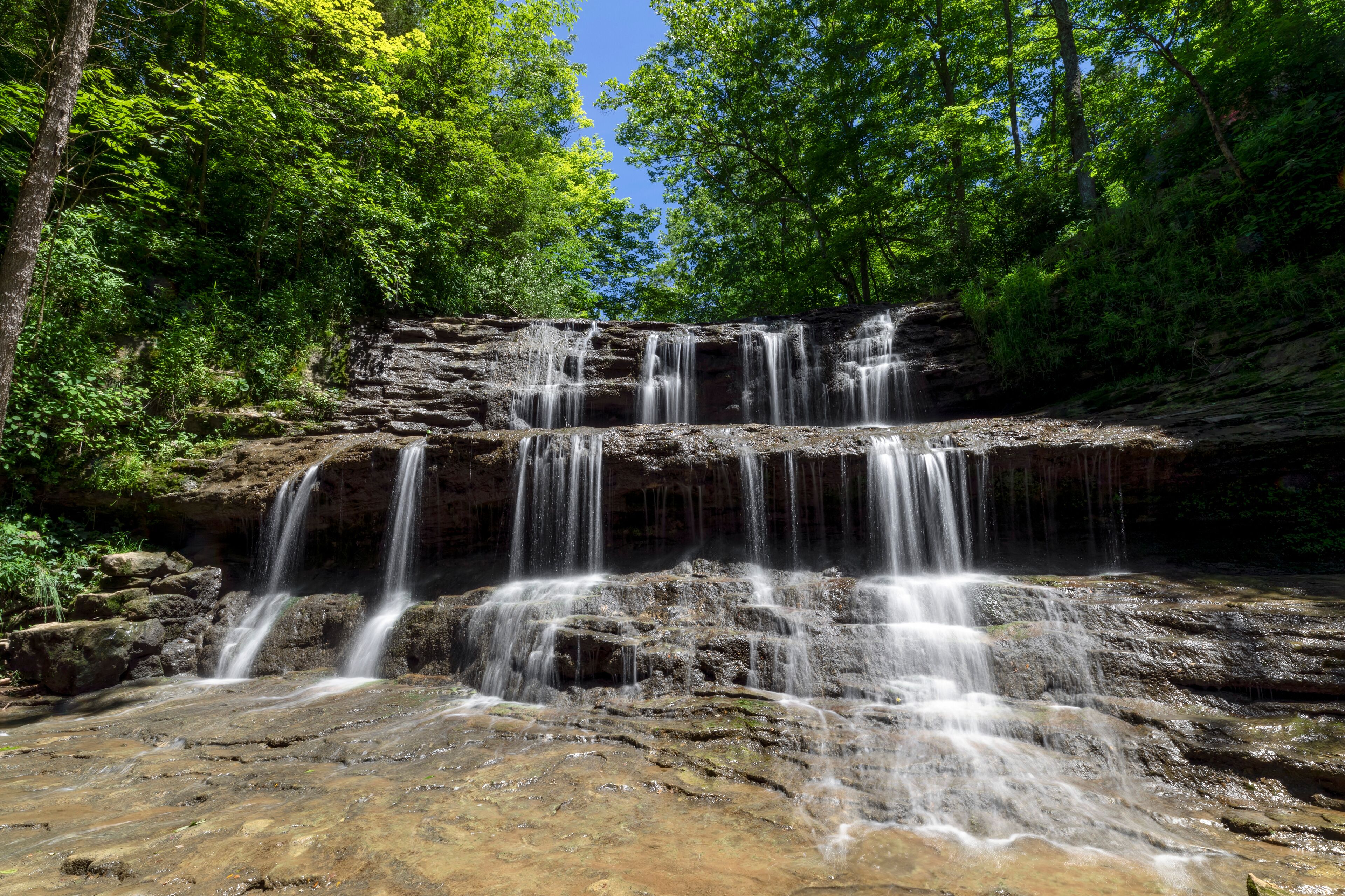 Rivulets of falling water over rock ledges are topped by emerald green leaves and a deep blue sky on a summer day at Fallsville Falls, a beautiful tiered waterfall near Hillsboro, Ohio.