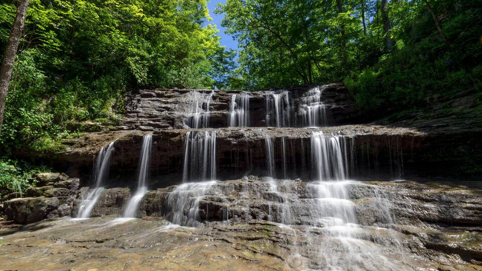 Rivulets of falling water over rock ledges are topped by emerald green leaves and a deep blue sky on a summer day at Fallsville Falls, a beautiful tiered waterfall near Hillsboro, Ohio.