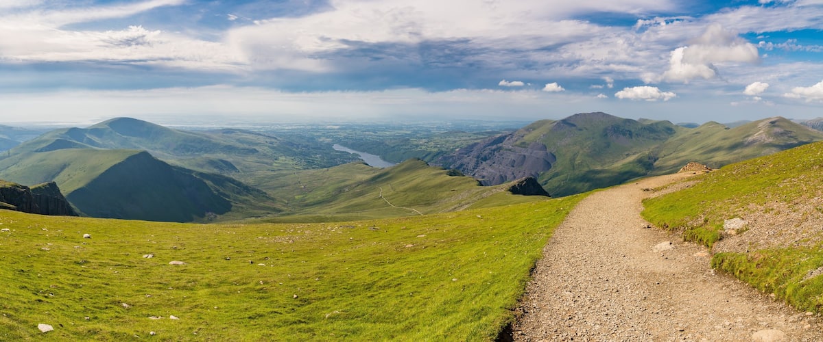A mountainous landscape with a gravel path, green hills, and a valley with a river.