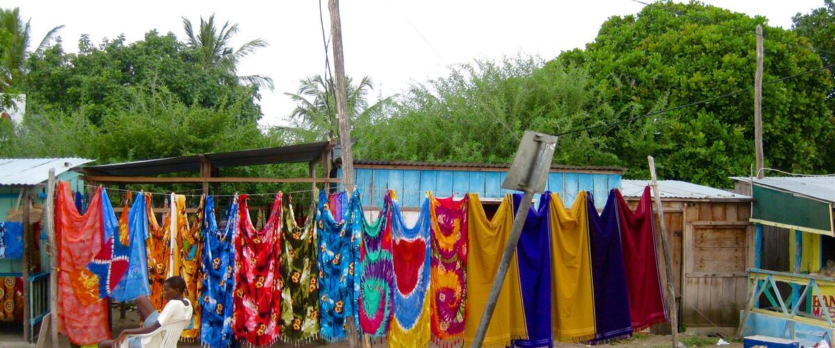 A rural setting with colorful fabrics hanging on a line, a person sitting on a plastic chair, and a small wooden structure in the background.