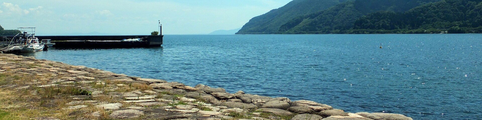 Una costa rocosa con un muelle y barcos, un lago tranquilo y montañas al fondo.