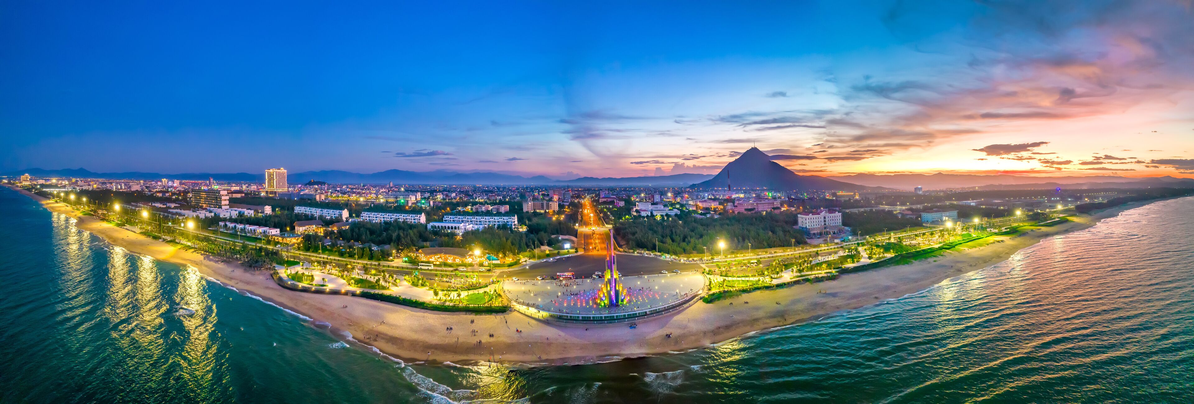 A coastal cityscape at sunset with a prominent mountain in the background.