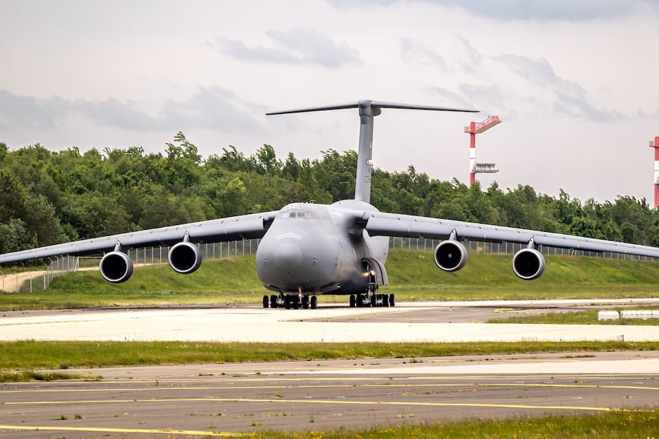 Un gran avión de carga militar en una pista con un bosque de fondo y dos grúas rojas.
