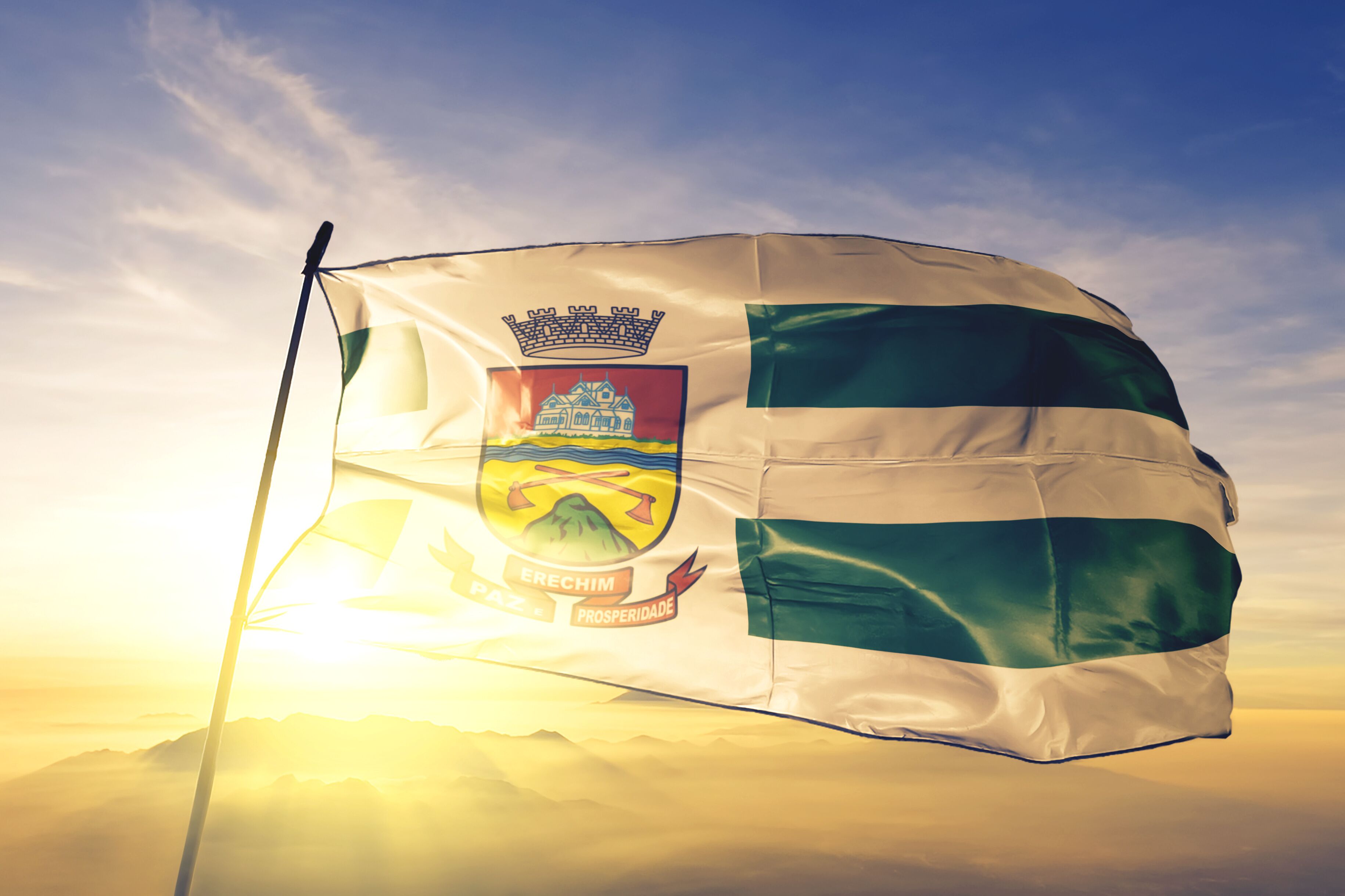 A flag with a coat of arms, a crown, and the words "Erechim" and "Prosperidade" on a flagpole against a sunrise sky.