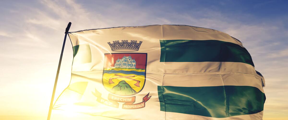 A flag with a coat of arms, a crown, and the words "Erechim" and "Prosperidade" on a flagpole against a sunrise sky.