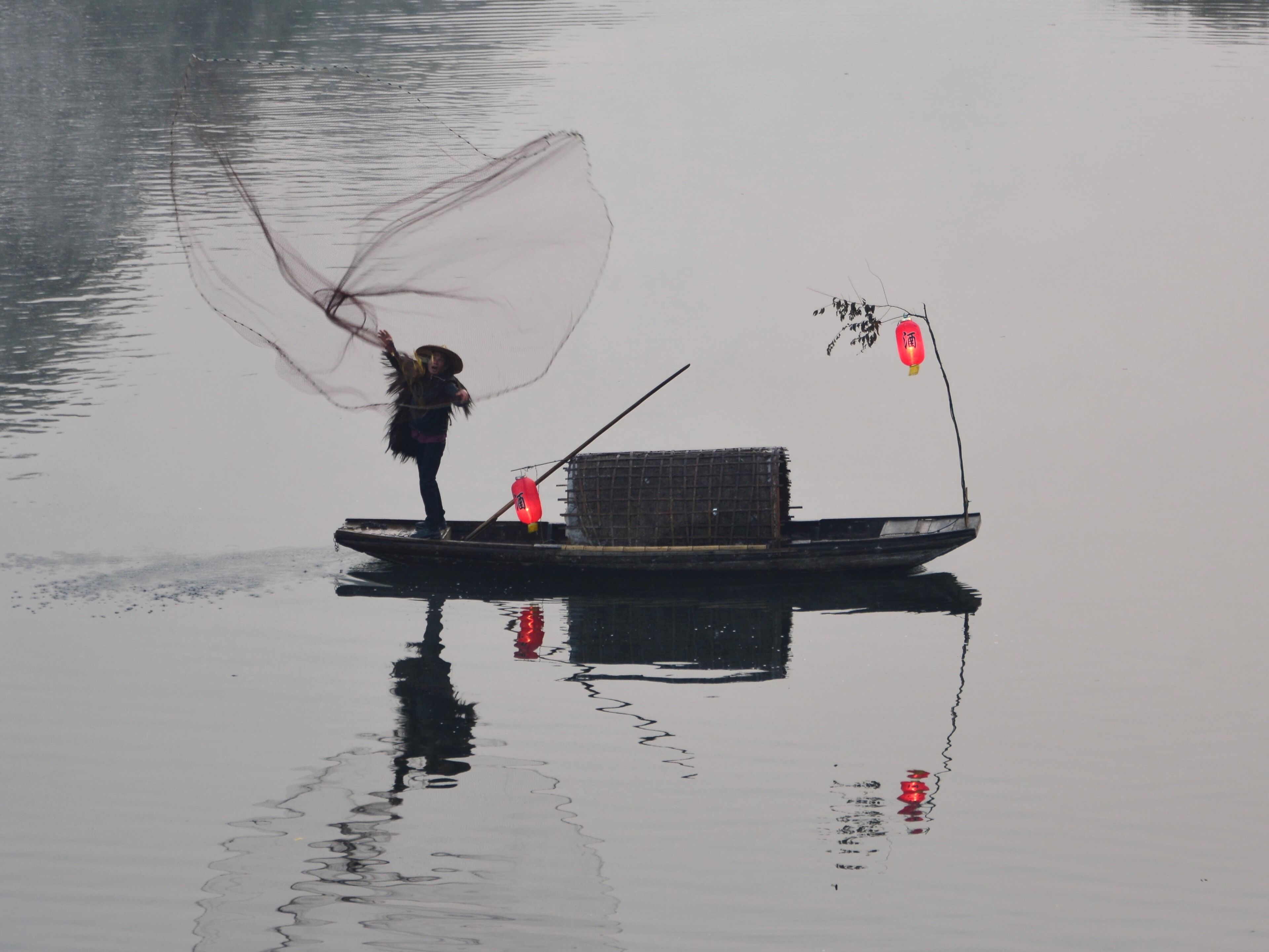 A person fishing on a small boat with a net, a lantern, and a cage.