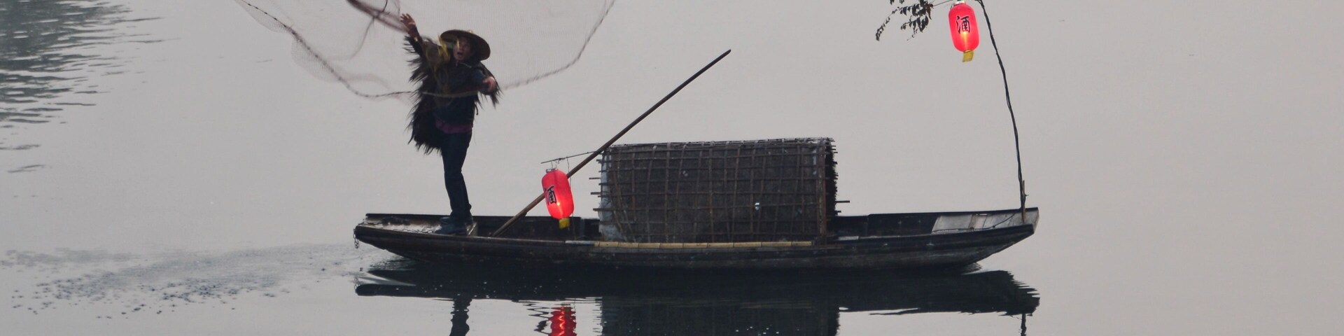 A person fishing on a small boat with a net, a lantern, and a cage.