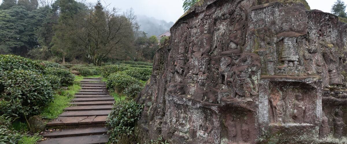 A stone pathway leading to a large rock with carvings, surrounded by greenery and trees.