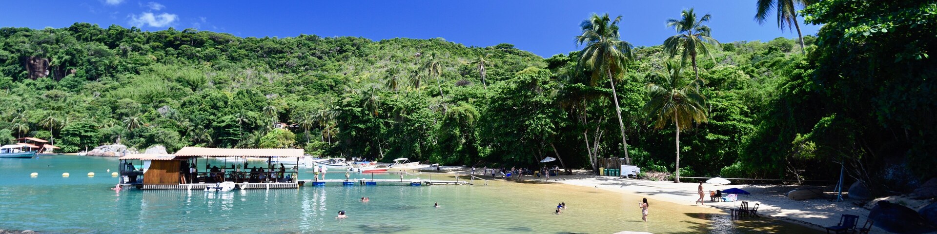 Una playa tropical con agua cristalina, rocas grandes, palmeras y un muelle.