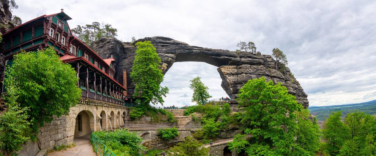 A historic building with a stone arch bridge, a large rock arch formation, and lush greenery.