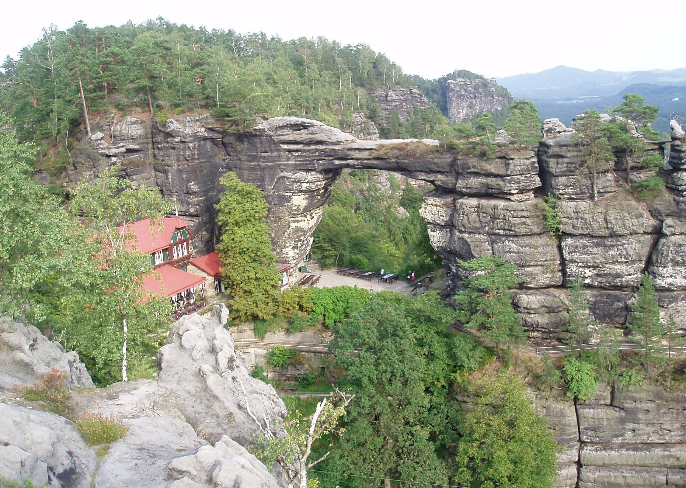 A natural rock arch formation with a red-roofed building and surrounding forest.