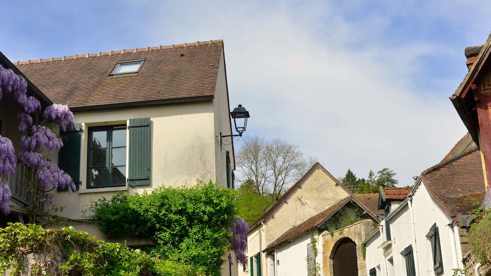 A quaint European village with traditional architecture, including a house with purple wisteria and a stone archway.
