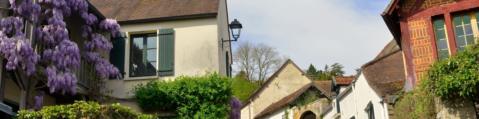 A quaint European village with traditional architecture, including a house with purple wisteria and a stone archway.