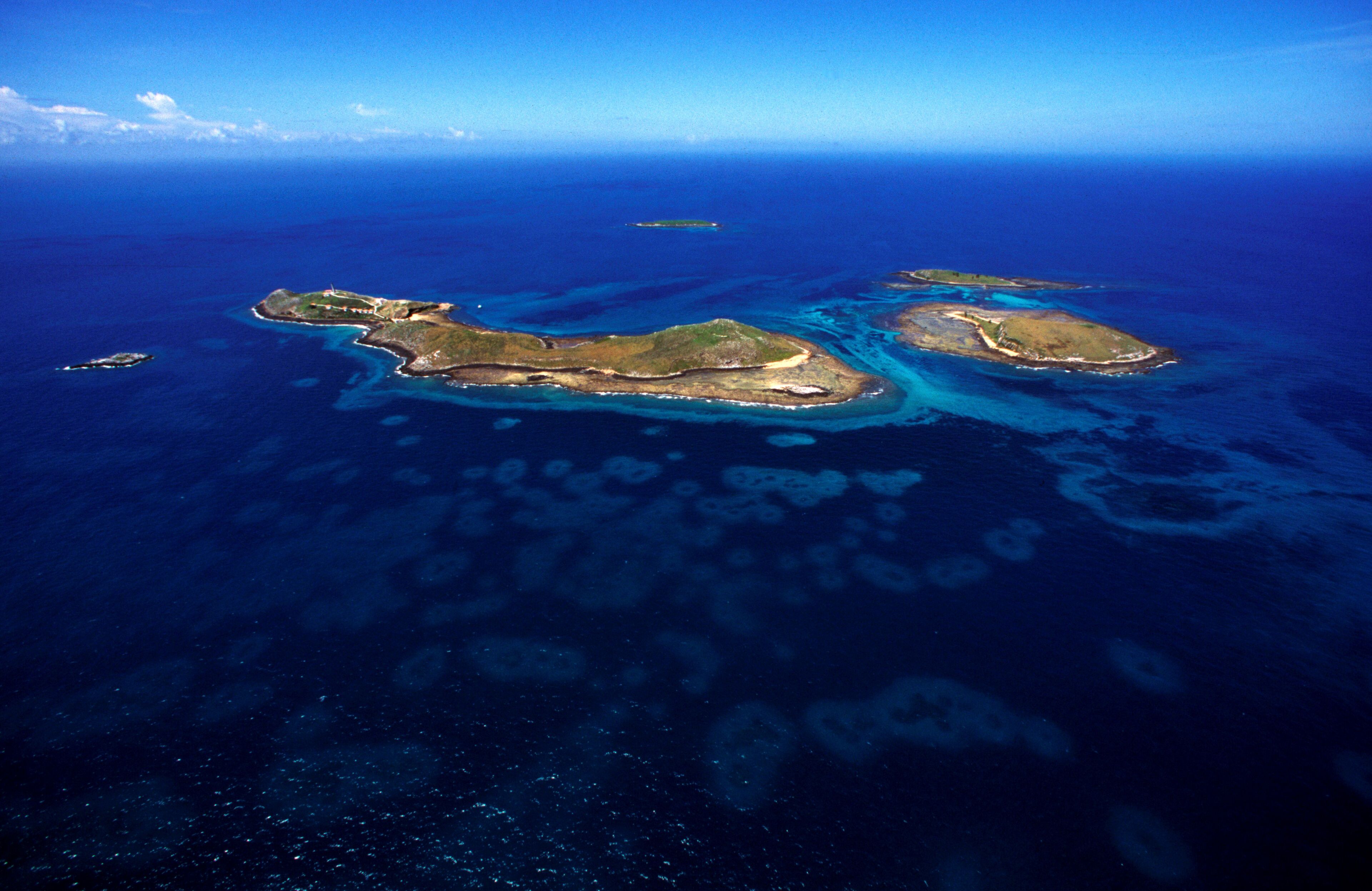 An aerial view of a chain of islands with clear blue waters and shallow coral reefs.