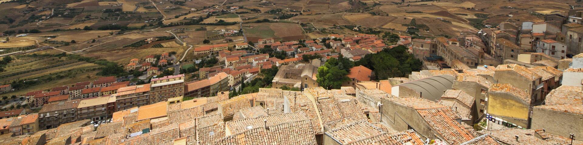 A panoramic view of a town with terracotta-roofed buildings, rolling hills, and a clear blue sky with scattered clouds.