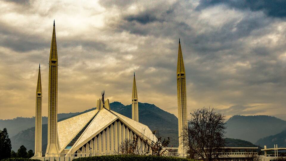 A mosque with four tall minarets and a large central dome, surrounded by mountains and trees.