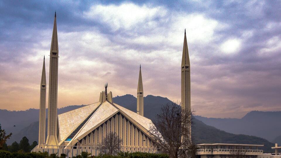 A mosque with four tall minarets and a large central dome, surrounded by mountains and a cloudy sky.