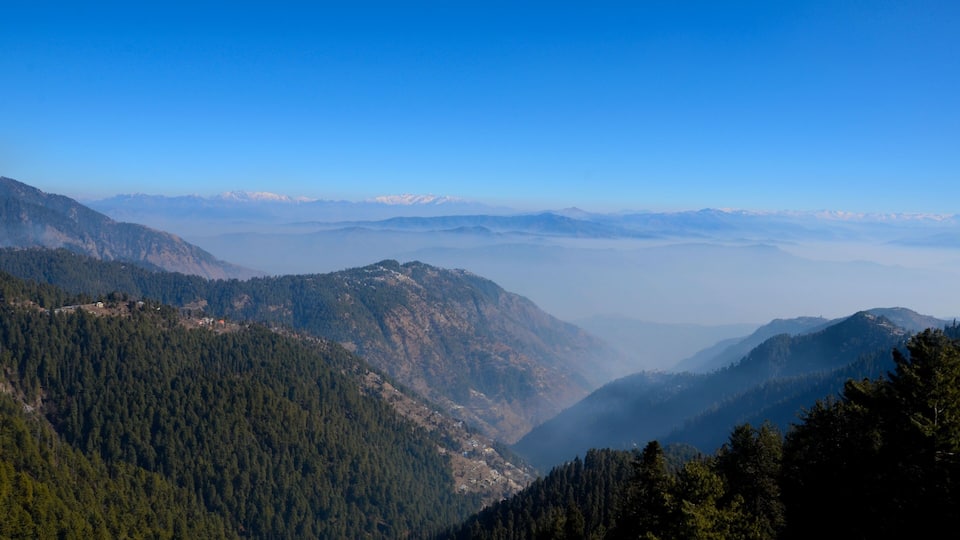 A mountainous landscape with dense forests, a valley, and distant snow-capped peaks under a clear blue sky.