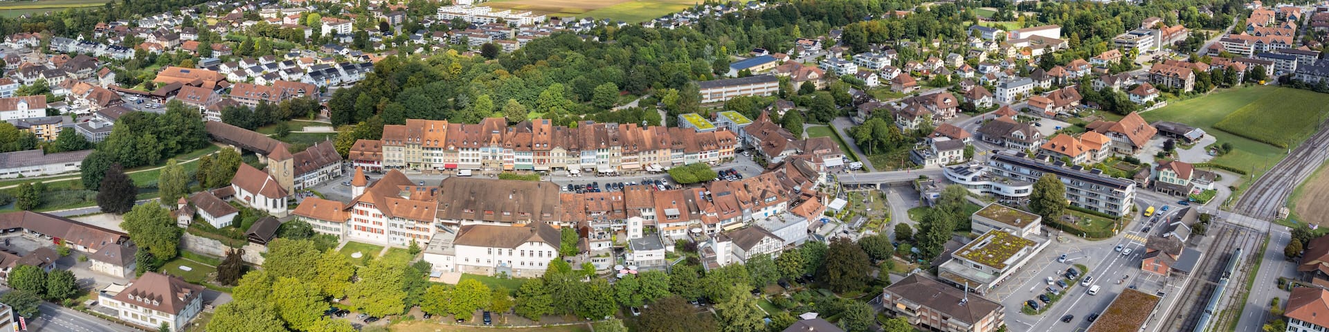 Aerial view of quaint rooftops cascading down towards the verdant fields, framed by the majestic mountains under a bright sky, Aarberg, Canton of Bern, Switzerland.