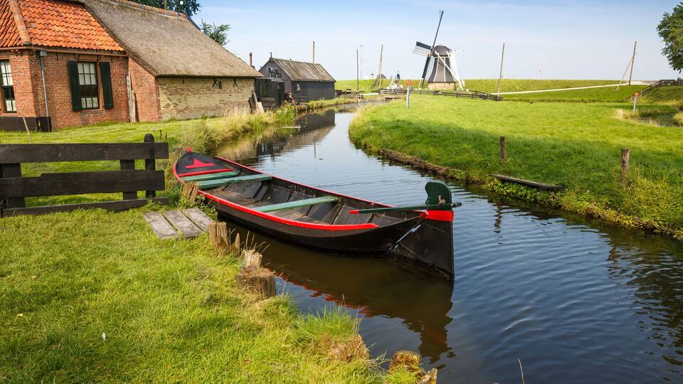 A narrow canal with a traditional boat docked on the left, a windmill in the background, and a thatched-roof building.