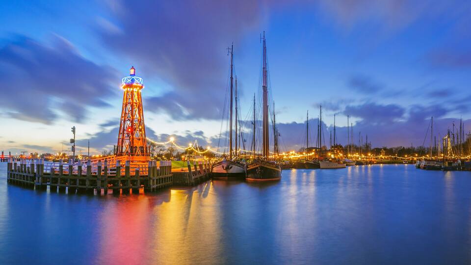A waterfront with a brightly lit tower, several sailboats, and a calm sea at dusk.