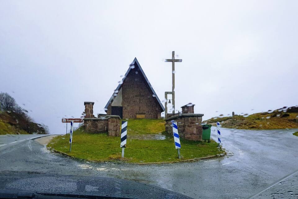 A small stone church with a bell tower and a cross, surrounded by grassy terrain and a wet road.
