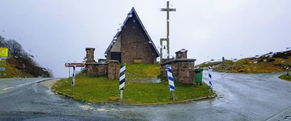 A small stone church with a bell tower and a cross, surrounded by grassy terrain and a wet road.