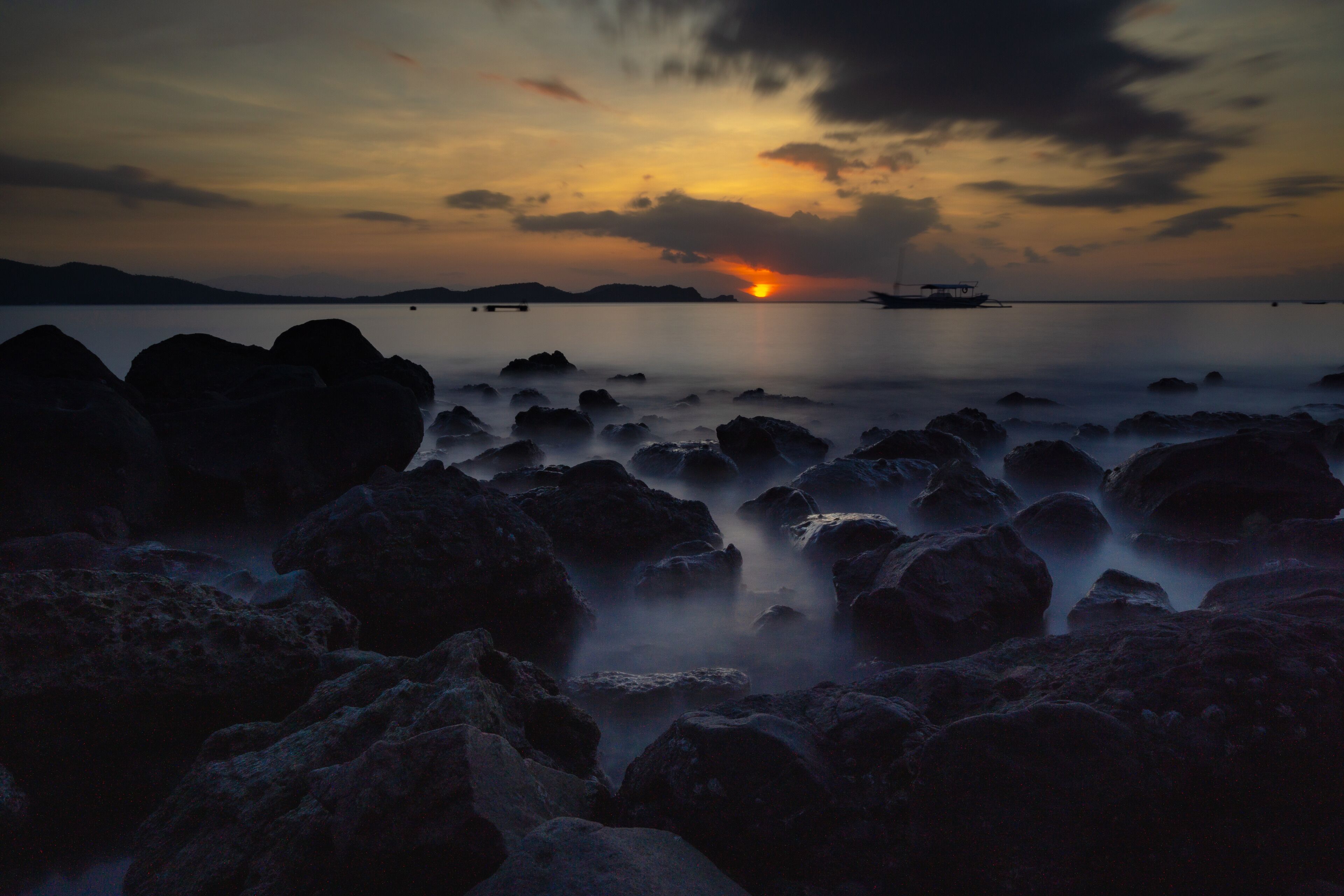 A sunset over a calm sea with rocky shores and a distant boat.