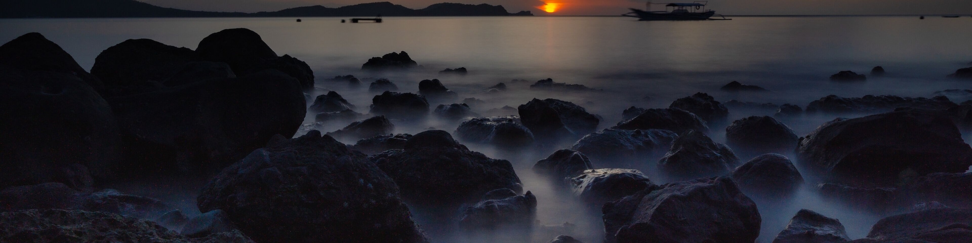 A sunset over a calm sea with rocky shores and a distant boat.