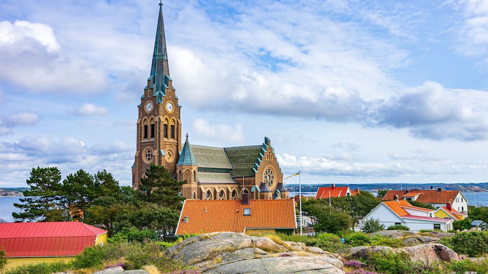 A church with a tall spire, a red-roofed building, and a body of water in the background.
