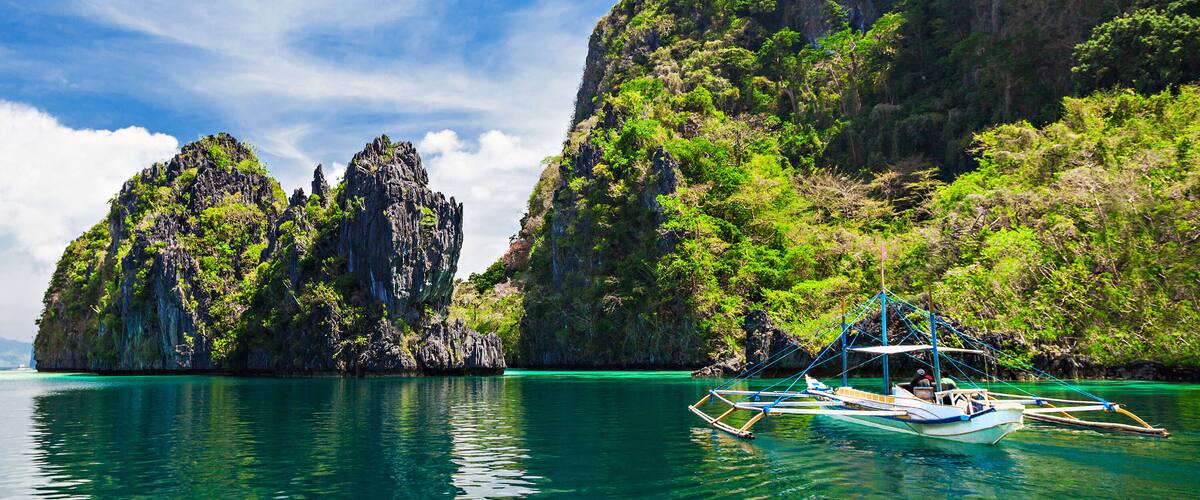 A clear turquoise sea with a traditional boat anchored near a rocky island with lush greenery.