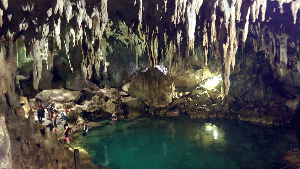 Une grotte ornée de stalactites et de stalagmites, une étendue d’eau et des personnes qui l’explorent.