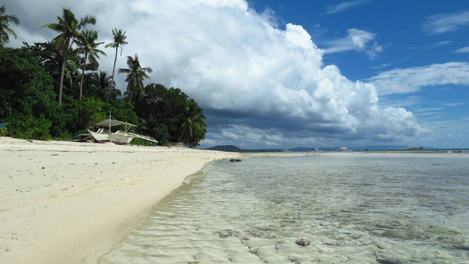 A tropical beach with clear shallow waters, palm trees, and a boat on the sand.