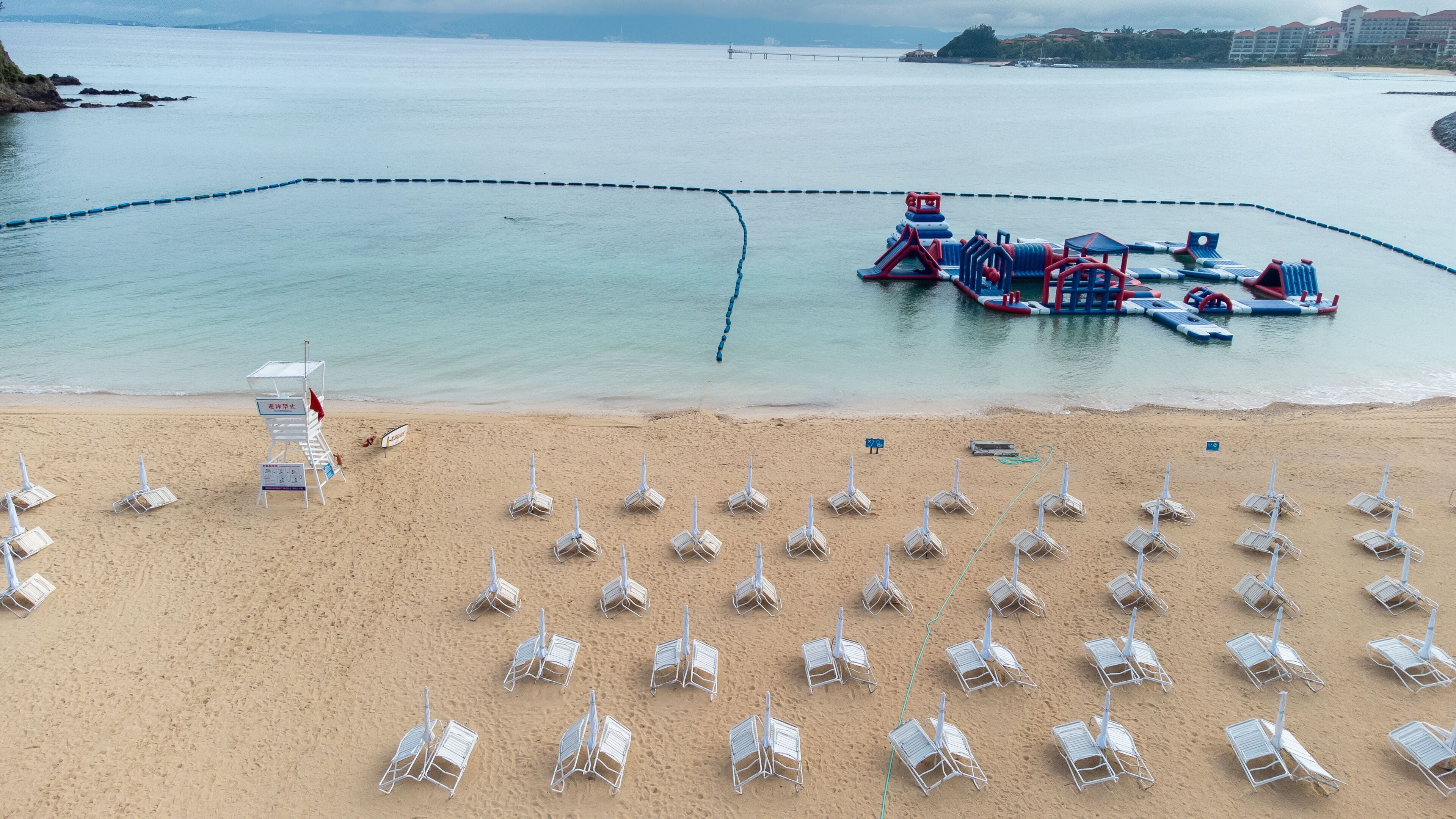 Uma praia com fileiras de espreguiçadeiras brancas e uma estrutura flutuante azul na água.