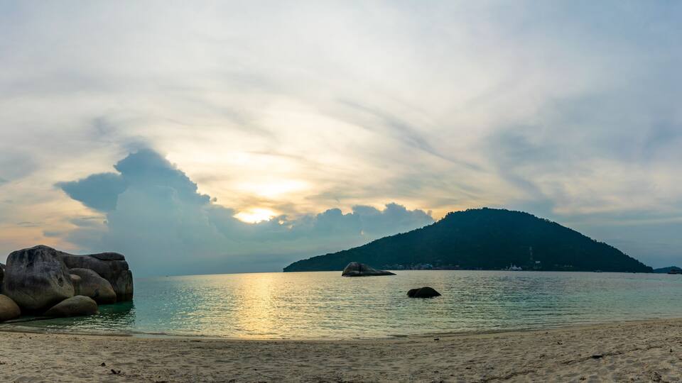 A beach with large rocks, calm water, and a mountain in the background.