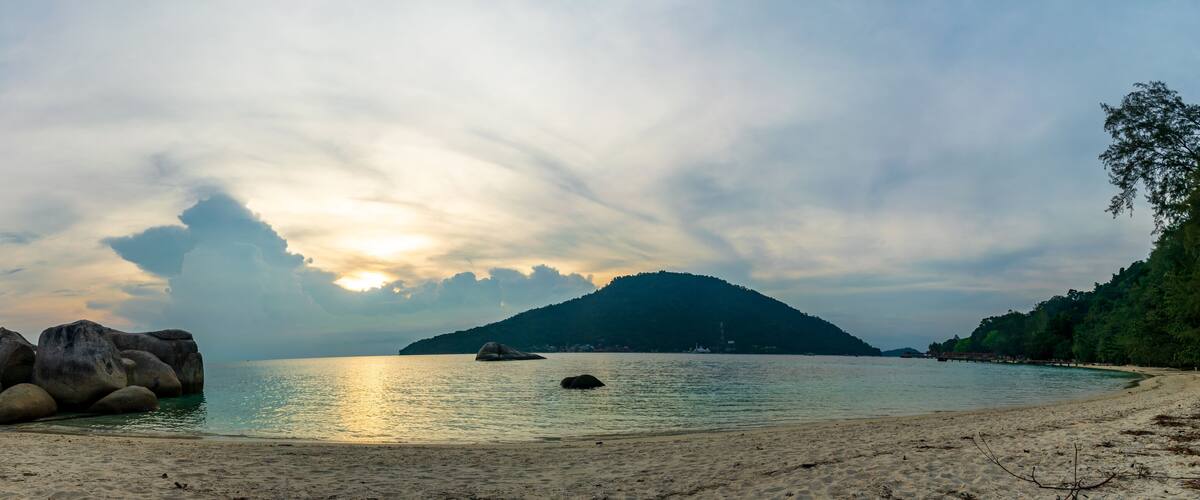 A beach with large rocks, calm water, and a mountain in the background.