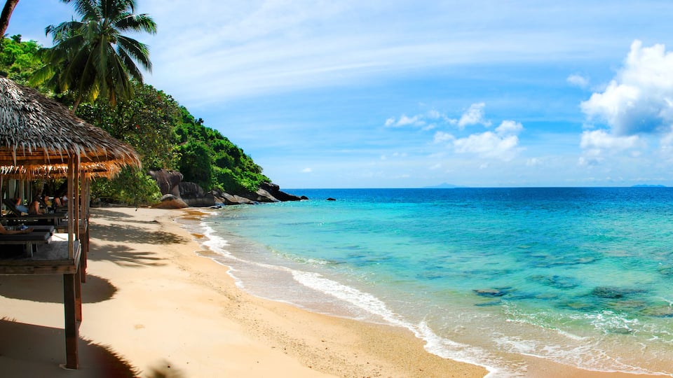 A beach with clear turquoise water, a thatched hut, and a pier.