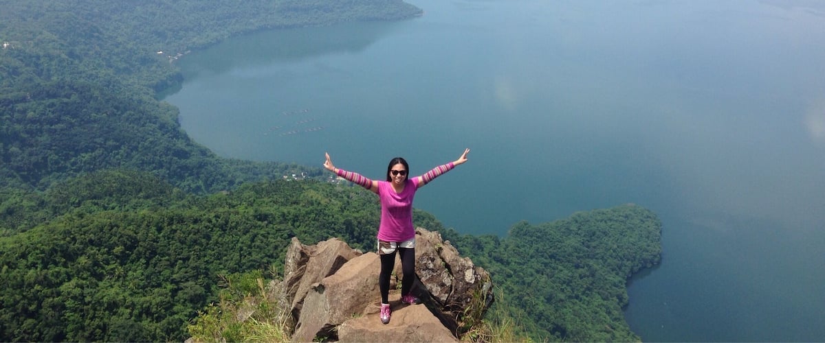 A person standing on a rocky outcrop with arms outstretched, overlooking a vast body of water surrounded by dense green forest.