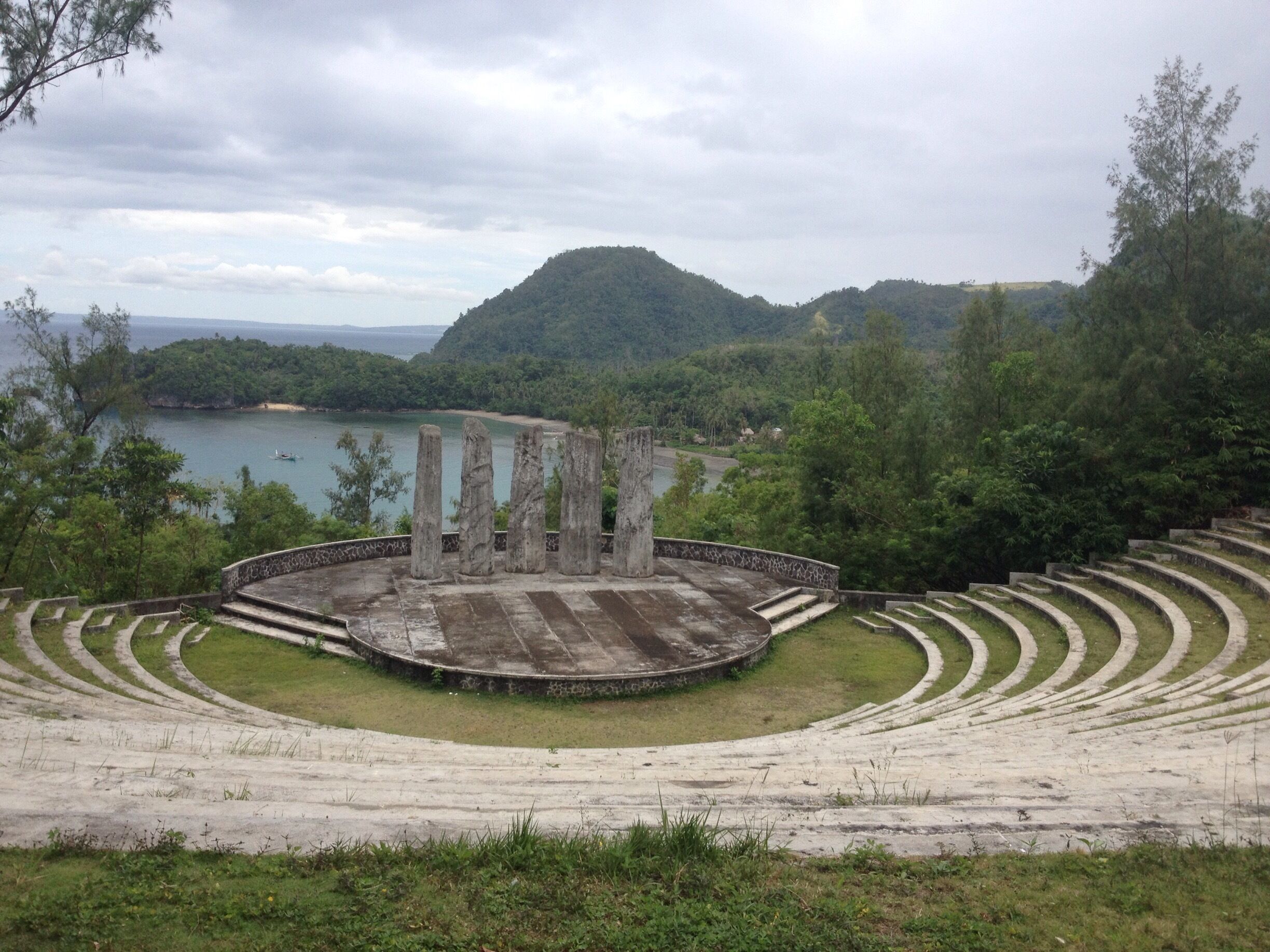 An amphitheater with a circular stage surrounded by tiered seating, overlooking a lake and forested hills.