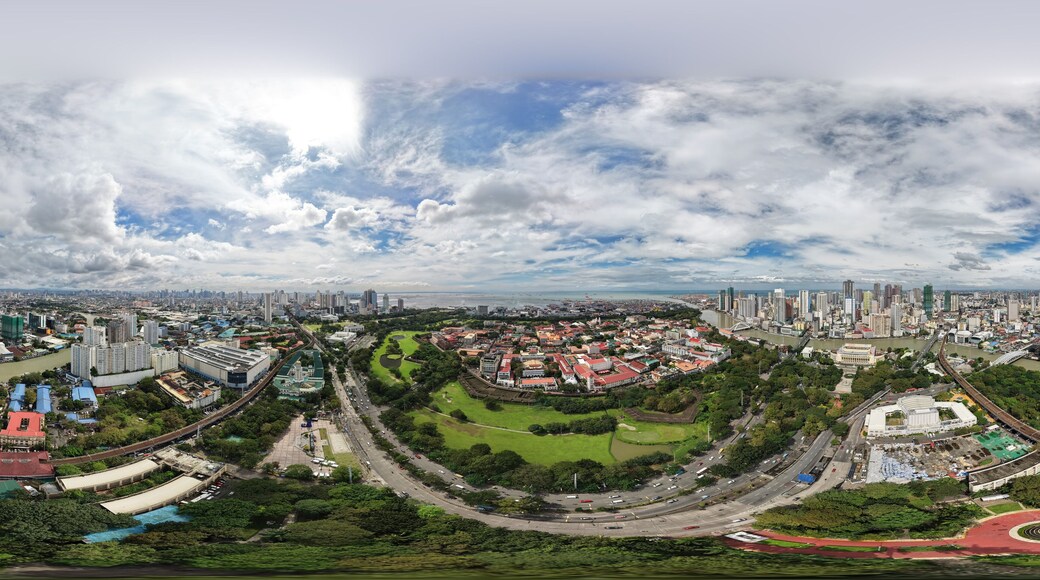 High altitude 360 aerial panorama of Intramuros historic district in Manila, Philippines, with golf course, green parks, Pasig River and city skyline under dramatic cloudy sky.