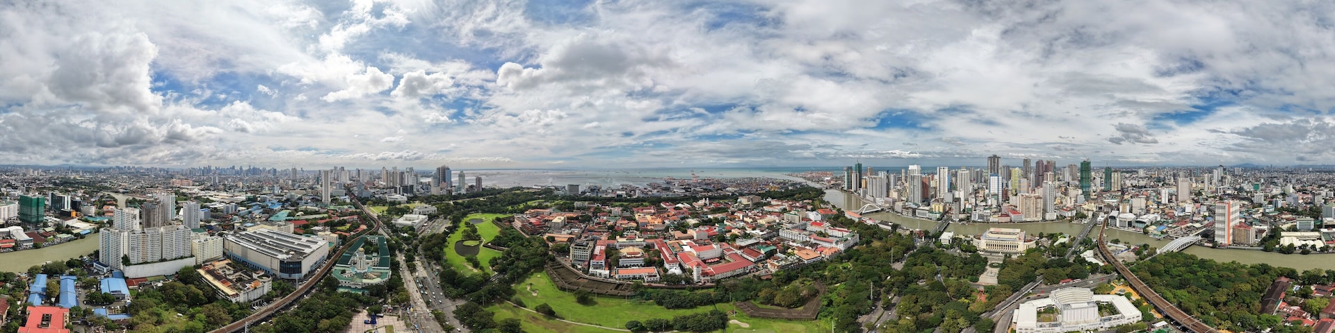 High altitude 360 aerial panorama of Intramuros historic district in Manila, Philippines, with golf course, green parks, Pasig River and city skyline under dramatic cloudy sky.
