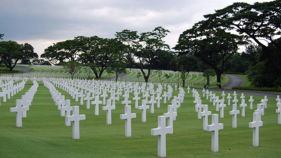A cemetery with rows of white crosses on green grass, surrounded by trees and a winding road.