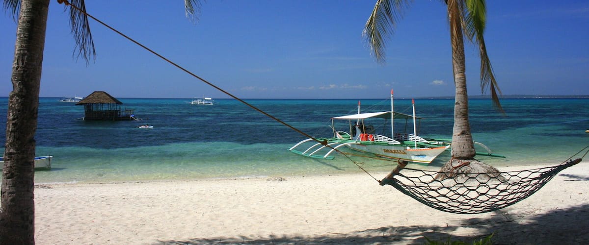 A hammock between palm trees on a beach with clear turquoise water and boats in the distance.