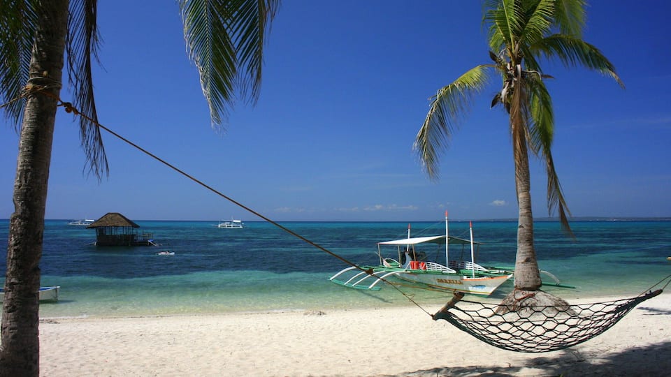 A hammock between palm trees on a beach with clear turquoise water and boats in the distance.